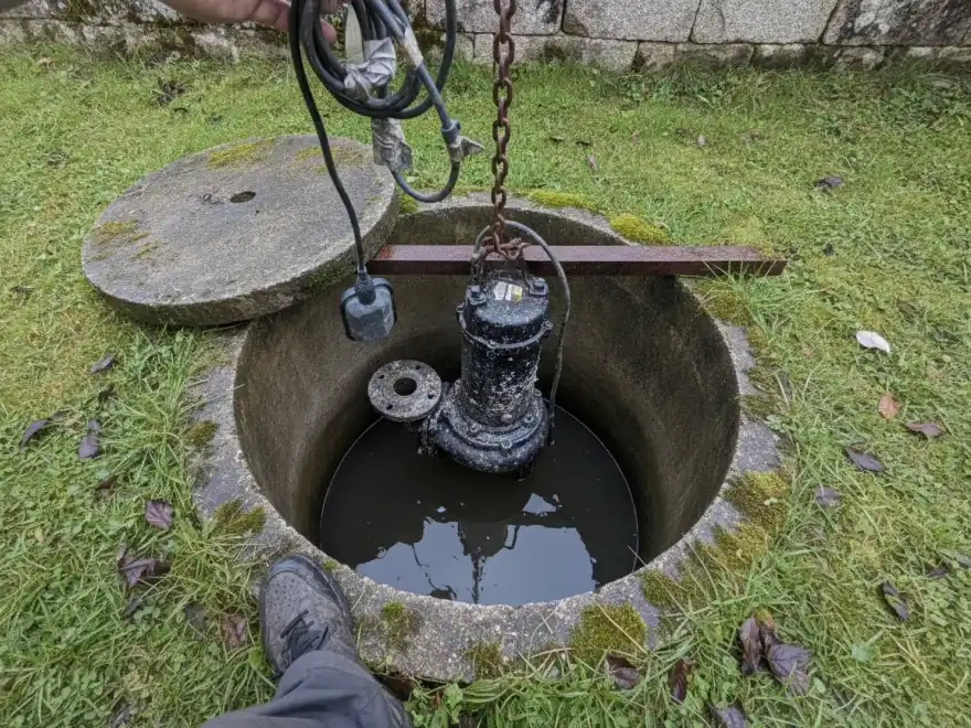 Pompe de relevage eaux usées à Sarzeau : regard béton moussu ouvert dans une pelouse bretonne, pompe encrassée sur sa chaîne (illustration)